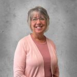 Smiling woman with short gray hair and glasses wearing a pink cardigan, standing against a gray textured background.