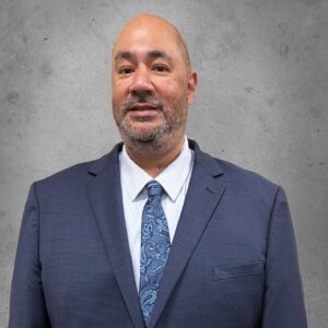 Professional portrait of a man in a navy suit and patterned blue tie standing against a gray wall background.